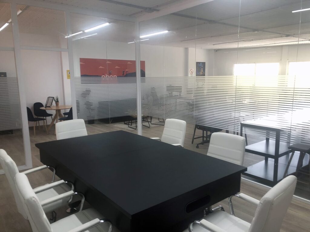 Un bureau de coworking moderne, aux murs de verre, situé à Coín, Málaga, Espagne. L'espace comprend une longue table noire et des chaises blanches, est bien éclairé par la lumière naturelle et présente des bandes givrées sur le verre pour plus d'intimité.