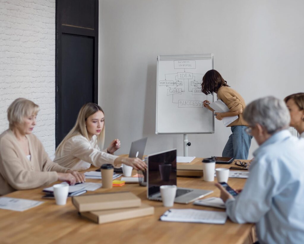 Coworking en Guadalhorce 2 En una oficina de coworking en Coín, Málaga, España, cuatro personas están sentadas en una mesa con ordenadores portátiles y papeles, trabajando en colaboración mientras una persona está de pie ante una pizarra blanca, ilustrando un diagrama.