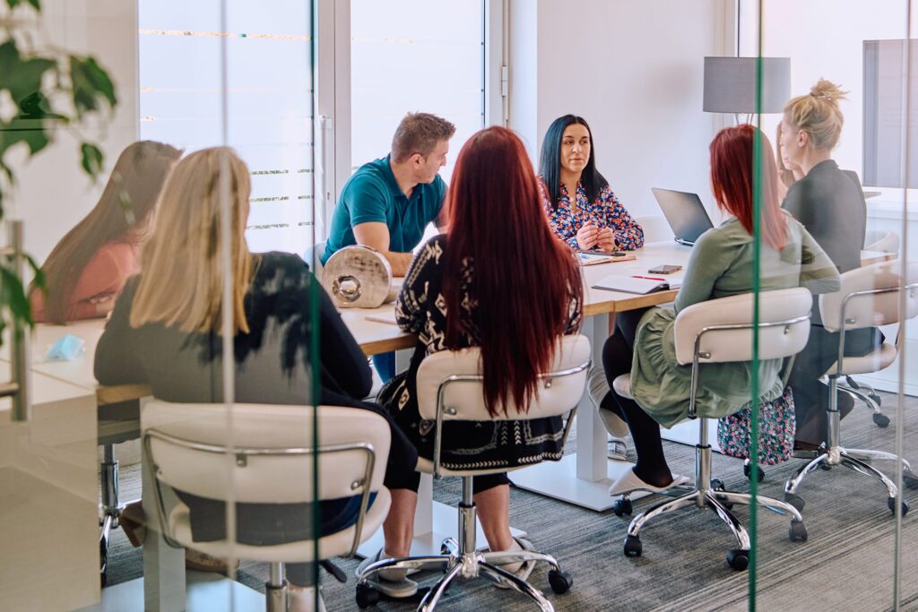Un groupe de six personnes est assis autour d'une table de réunion, en pleine discussion, dans un espace de coworking moderne doté de grandes baies vitrées et de chaises blanches, situé à Coín, près de Málaga.